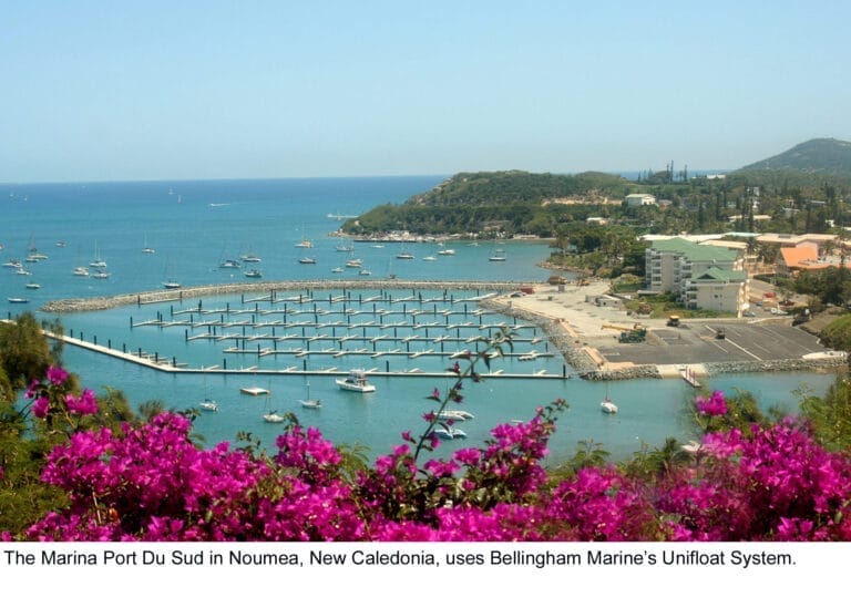 Noumea marina wave attenuator with boats docked, surrounded by clear blue water and lush green hills in the background.